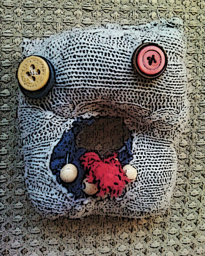 Image of a hand sewn momu monster with button eyes and a grey square body with a mouth made of beads and a cloth tongue sticking out.
